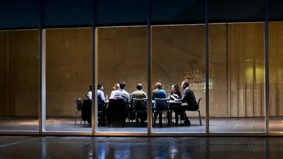 A team meeting at a long table, seen at night through a lit glass facade