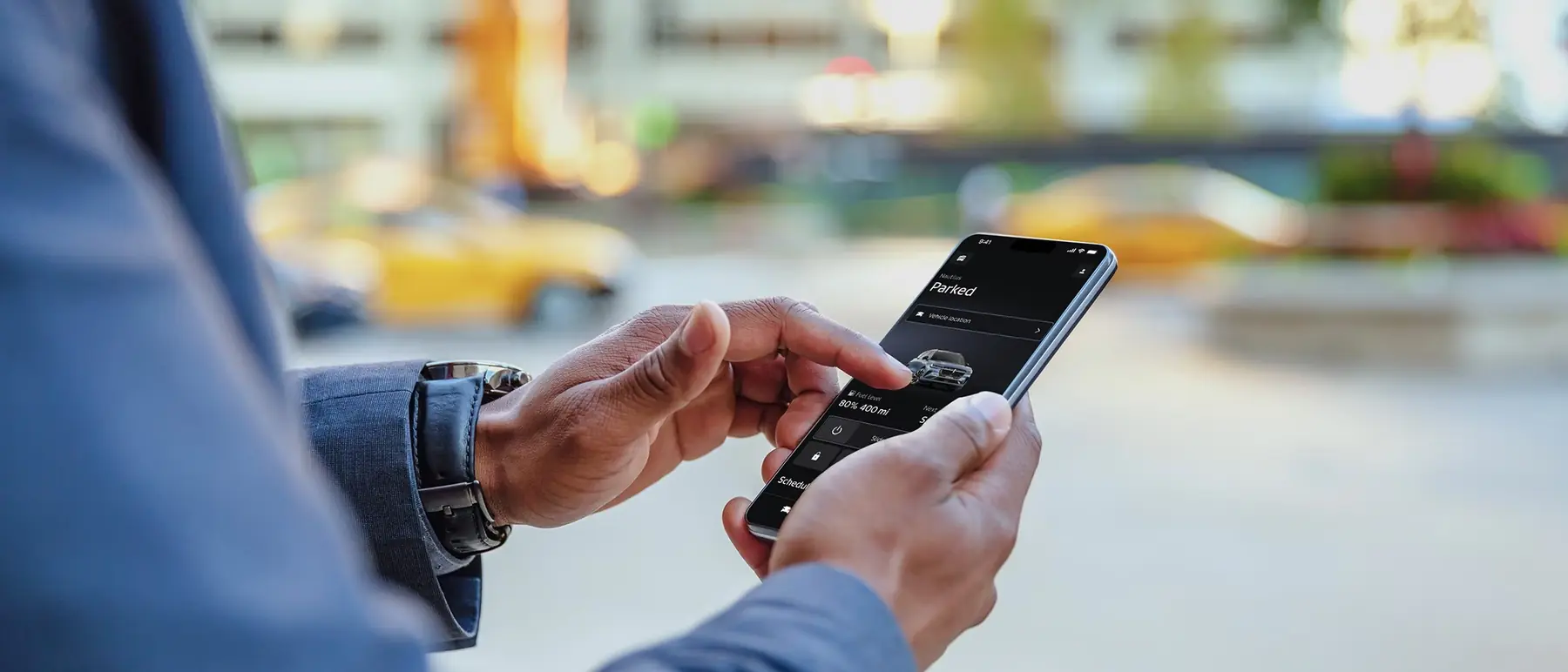 A person checking a connected vehicle app on a phone in the street, the car shown parked with charge remaining